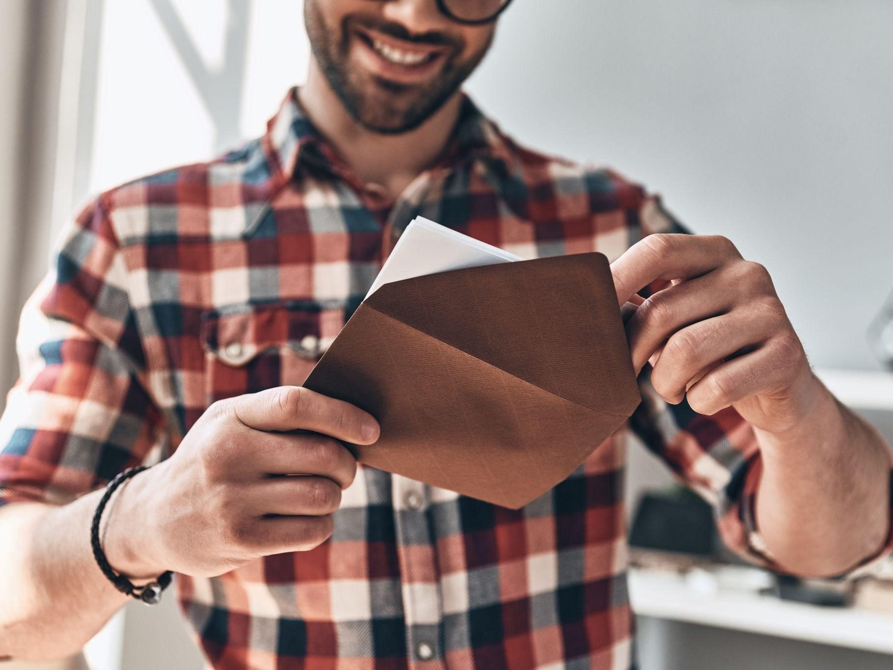 Young man with beard wearing a checked shirt smiling and opening direct mail in a brown envelope